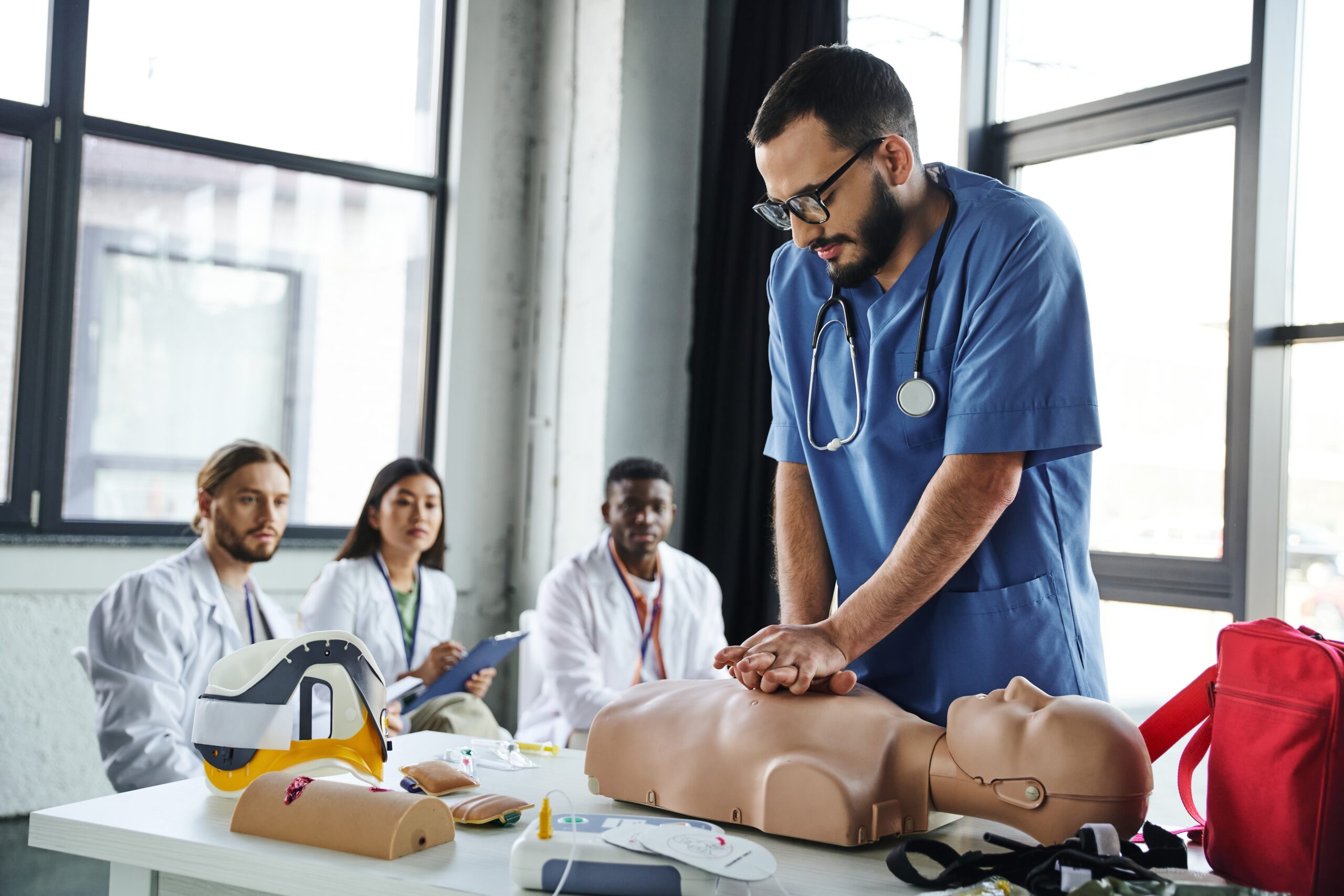 A male medical student with a beard and glasses, wearing blue scrubs and a stethoscope, performs chest compressions on a CPR mannequin.