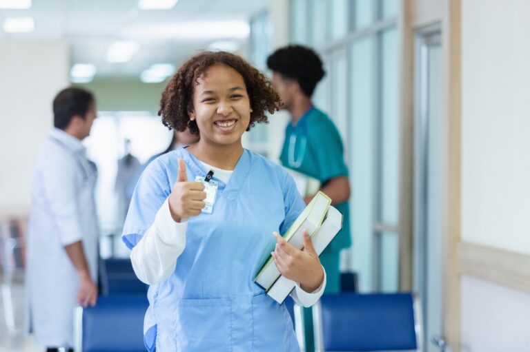 A young nursing student with curly brown hair stands in a brightly lit hospital hallway, smiling broadly and giving a thumbs-up. She is wearing blue medical scrubs over a white long-sleeved shirt, has an ID badge clipped to her collar, and is carrying two textbooks.