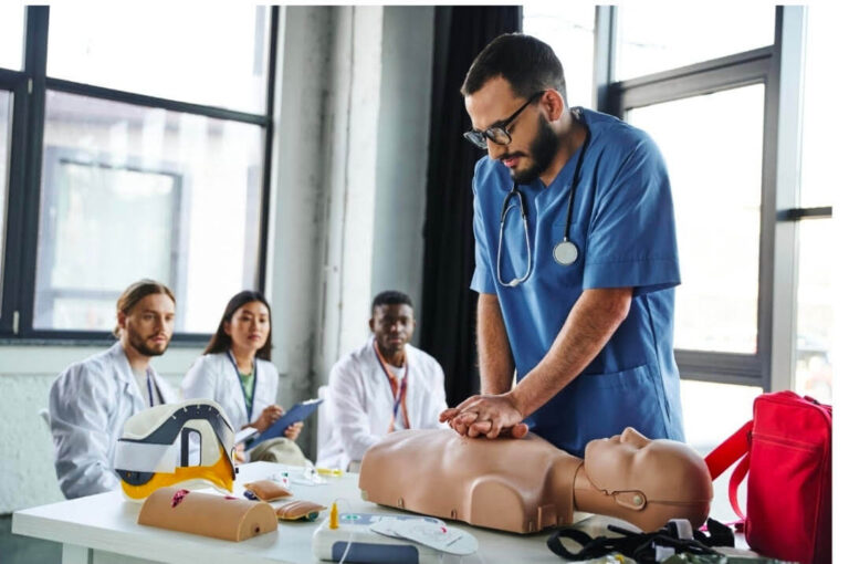 A medical student with a beard, glasses, and blue scrubs performs chest compressions on a CPR training mannequin in a bright classroom setting.