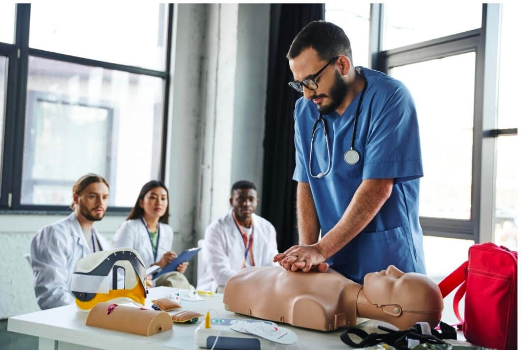 A medical student with a beard, glasses, and blue scrubs performs chest compressions on a CPR training mannequin in a bright classroom setting.